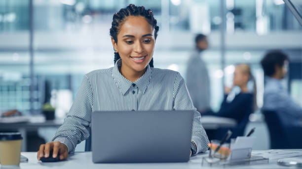 istock Multi-Ethnic Office: Professional Black Female IT Engineer Working on Desktop on Laptop Computer. Successful, Smiling Businesswoman with Braided Hair Does Creative Job. Confident Product Designer 1364004774