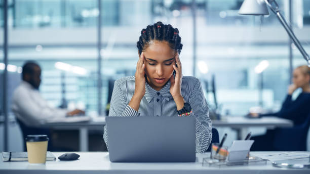 young black woman office worker uses laptop, feels sudden burst of pain, headache, migraine. overworked accountant feeling project pressure, stress, massages her head, temples. front view portrait - frustração imagens e fotografias de stock