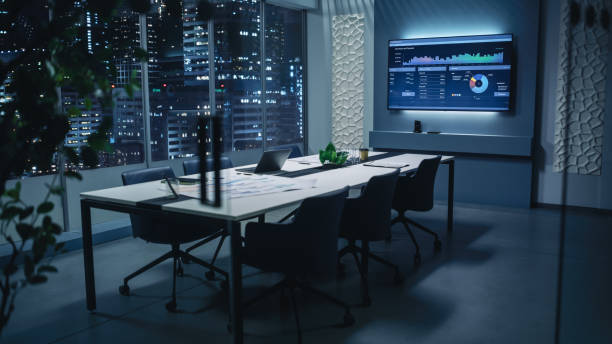 Modern Empty Meeting Room with Big Conference Table with Various Documents and Laptop on it. Wall TV Showing Company's Growth, Statistics, Graphs and Pie Charts. Late Evening or Night City Outside.