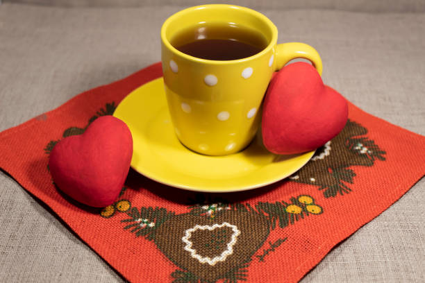 A cup of tea on a yellow saucer stands on a red napkin. Two hearts lie on the sides.