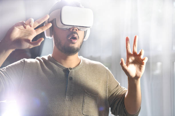istock Young Asian man wearing VR goggles while playing video games with hands reaching out to touch something in virtual world 1364016504