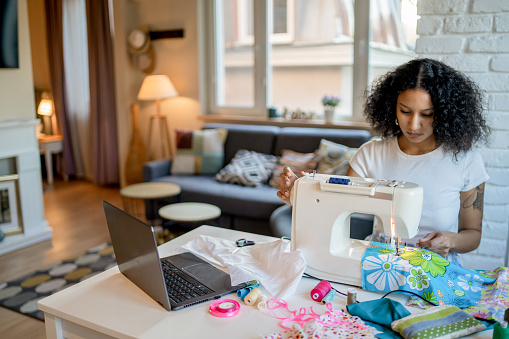 A seamstress is creating homemade items to sell on her online business, with her laptop handy to keep an eye on new orders
