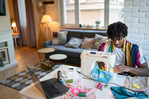 Looking down at a dining table in a modern apartment, a multi-racial man is sewing fabric on a sewing machine, a laptop open on the table surrounded by sewing threads, ribbons and material