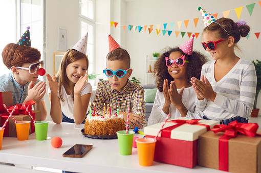 Group of children in funny party hats and sunglasses celebrating their friend's birthday. Little girl blowing candles on cake and making a wish surrounded by happy diverse classmates clapping hands