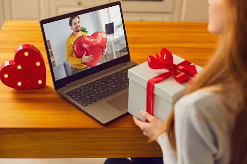 Over shoulder close-up virtual date of happy couple on Saint Valentine's Day in lockdown. Young man and woman in love showing presents they prepared. Girl holding gift box and video calling boyfriend