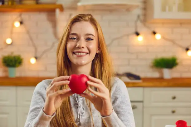 Video call headshot of happy smiling young woman holding red heart. Concept of charity and care, donation, sharing love and kindness, saying thank you or giving presents for Saint Valentine's Day