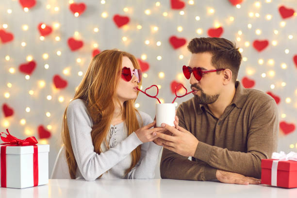 Young couple sitting at table in cafe on Saint Valentine's Day and sipping drink through straws Young man and woman having a fun date on Saint Valentine's Day, sitting at table in cafe or at home, sipping drink from one cup through heart-shaped straws and enjoying cute and funny couple moment drinking straw funny stock pictures, royalty-free photos & images