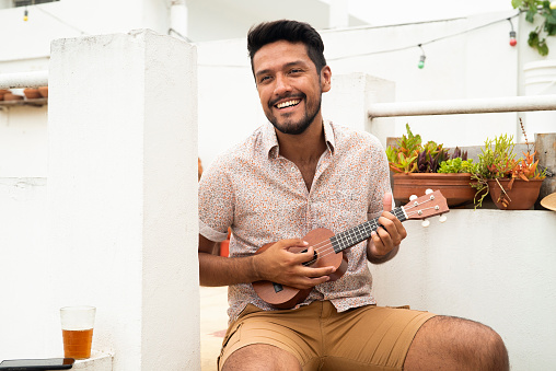 Portrait of young adult latin man having fun while playing ukulele in party while sitting outdoors