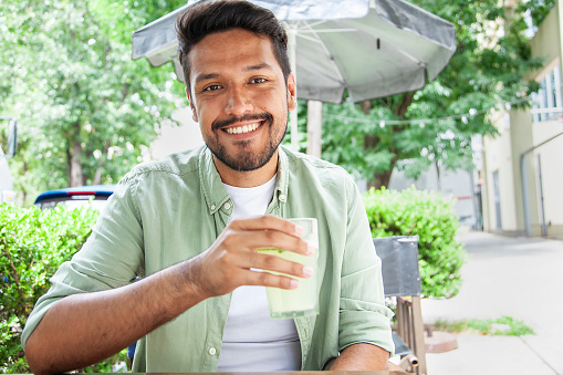 Front shot of cheerful young mexican man looking at the camera while drinking lemonade sitting on chair in sidewalk