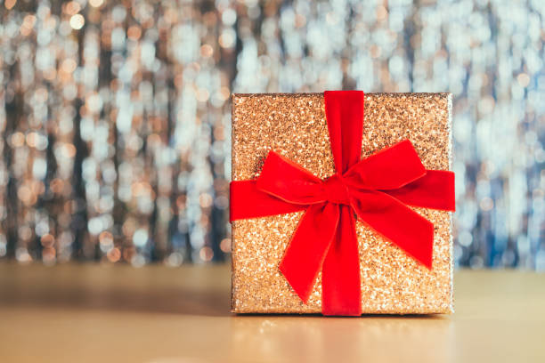 сверкающая золотая подарочная коробка с бархатно-красной лентой с бантом на сверкающем золотом фоне мишуры. концепция подарка на день рожд - bow christmas red velvet стоковые фото и изображения