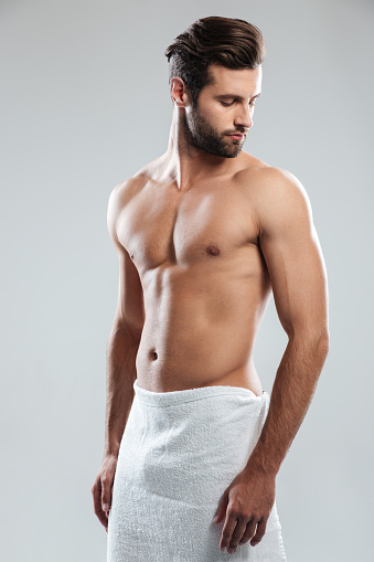 Photo of handsome young man dressed in towel standing isolated over grey background. Looking aside.
