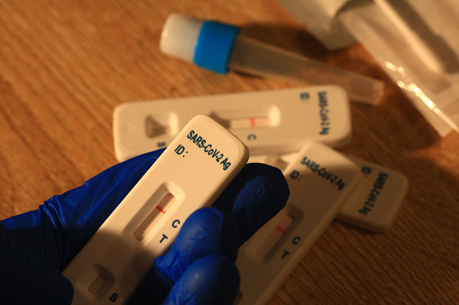 It's a hand wearing a blue glove and holding a negative covid 19 (sars-cov-2) self-test in the hand, and with other self-test and test kit items in the background