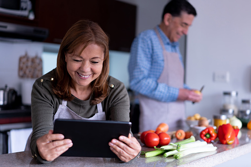 Happy Latin American couple preparing a meal together, mature woman looking for an online recipe on tablet while husband peels potatoes at background - Lifestyles