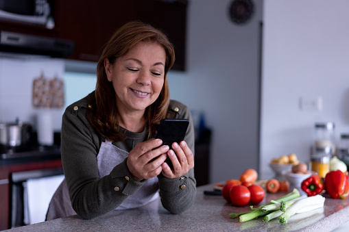 Cheerful Latin American woman texting on her smartphone while leaning on kitchen counter before cooking a meal at home - Lifestyles