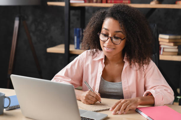 istock Young african tutor freelancer student schoolgirl writing homework at copybook, doing assignment on laptop remotely online, e-learning concept. 1364072597