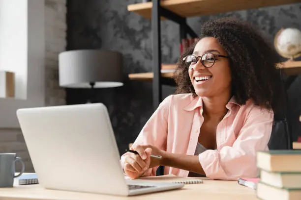 Cheerful laughing young african woman tutor freelancer student writing at copybook homework test, e-learning, working remotely, watching webinar on laptop