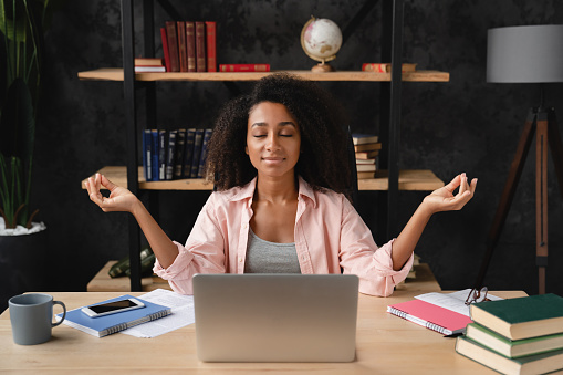 Young african freelancer worker businesswoman student meditating at workplace desk after working hard with homework, e-learning, feeling overworked and stressed