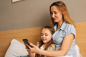 Happy young mother hugging to preschool daughter together browsing and surfing internet on mobile phone, chatting on video call
