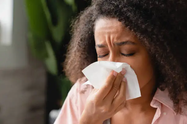 Photo of Sneezing coughing ill young african woman using paper napkin, having runny nose, blowing her nose. Coronavirus, infectious disease, flu, cold.