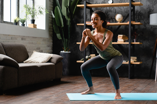 African young slim sportive woman teenage girl doing squats, stretching her body, legs at home on fitness mat. Training workout on isolation.