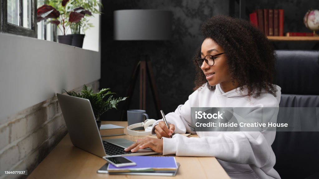 African young teenager e-learning on distance, using laptop, writing homework assignment in copybook, watching webinars online from home. University Student Stock Photo