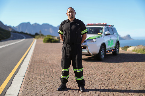 A mixed-race male paramedic wearing a protective face mask standing in front of his emergency response vehicle with emergency lights flashing..