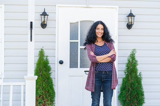 Curly smiling woman with arms crossed, standing in front of her house with white front door.