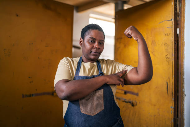 portret pewnej siebie afro kobiety noszącej ubranie robocze i wyglądającej na silną podczas zginania bicepsów - bicep women flexing muscles african descent zdjęcia i obrazy z banku zdjęć