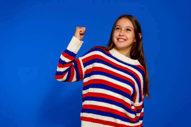funny little cute girl 10-11 years old wears a striped sweater making the gesture of winner say yes isolated on a pastel blue background child studio portrait. mother's day love family people lifestyle concept. - 10 11 years child human face female imagens e fotografias de stock