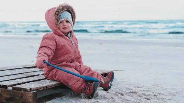 Adorable Caucasian Toddler Baby Girl Wearing Warm Winter Jumpsuit Playing in Seashore Beach Sand