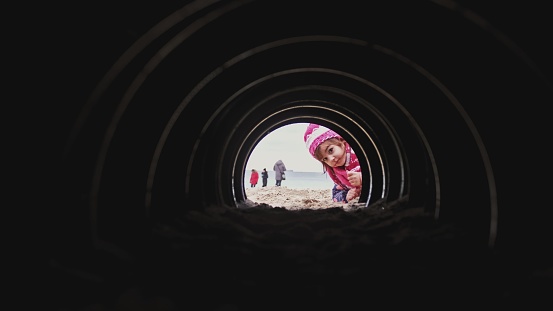Curious Caucasian Girl in Warm Clothes Looking and Crawling into Dark Narrow Tunnel on Beach