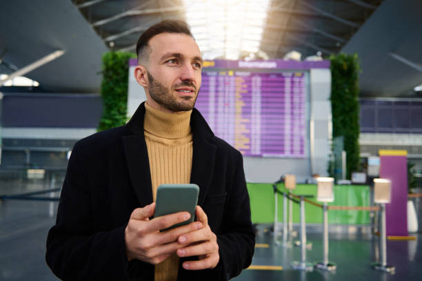 istock Handsome Caucasian young man using smartphone and checking flight standing in front of check-in board at departure terminal of international airport 1364089467