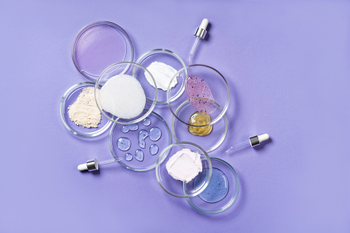 Cosmetic products, scrub, face serum and gel in many petri dishes on a pink background. Cosmetics laboratory research concept. Pastel violet background