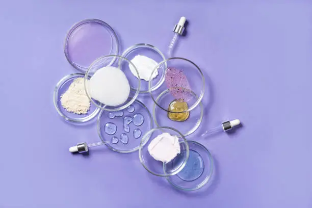 Photo of Cosmetic products, scrub, face serum and gel in many petri dishes on a pink background. Cosmetics laboratory research concept. Pastel violet background