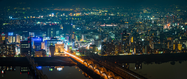 Panoramic view across the tranquil waters of the Yodo River surrounded by the crowded skyscraper cityscape of Osaka at night, Japan.