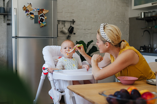 Caucasian blonde mother is feeding the baby in the kitchen. Baby (most probably a baby girl) is sitting in a feeding chair, looking at a camera and frowning while eating. The woman is holding a spoon and a  bowl is on a table behind her. She might be a single mother or a babysitter.