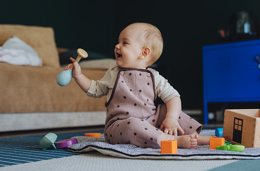 Laughing baby (either a boy or a girl) is sitting on the floor in a playroom and looking at a camera. There are different wooden toys on the floor around her. The kid is holding a toy.