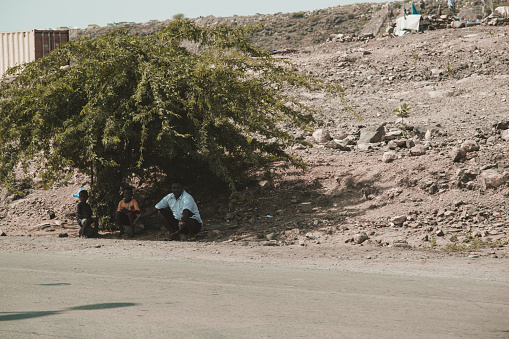 Djibouti, Djibouti - May 21, 2021: A Djiboutian man and his two sons sitting on the stone and waiting the bus under a tree's shade in Djibouti. Editorial shot in Djibouti