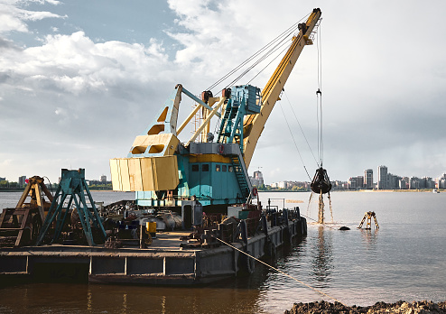 Industrial barge, working dredging crane near coast digging the ground, sunlight and dark clouds, reflection in calm river water