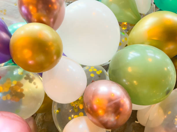 Close up of latex shiny balloons for party celebration – clear balloon with gold confetti inside, teal, purple, green, rose gold, and gold balloons all tight together stock photo