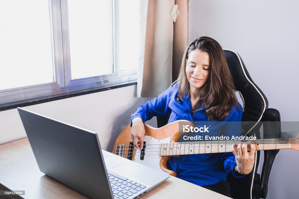 Woman studying music online in note book and playing electric bass guitar Woman wearing a blue shirt sitting at a table next to a window and with the help of her notebook watches online music class and plays her electric bass guitar 30-34 Years Stock Photo