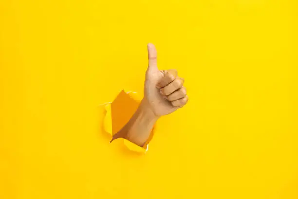 Photo of Close-up of male hand showing thumbs up on yellow background.