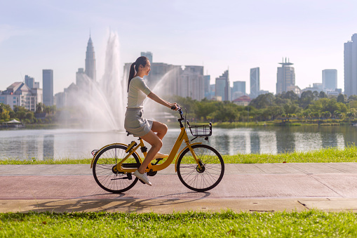 Asian woman uses bicycle sharing system, renting and riding road bike in an urban park for healthy exercise and relaxation with the city skyline in the background