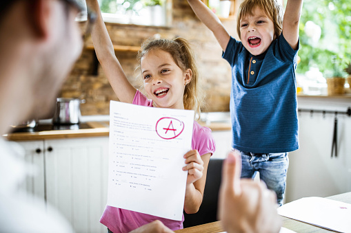 Happy little girl feeling satisfied while showing her father an A grade on her test. Her brother is celebrating in the background.