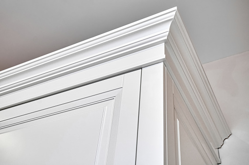 Crown moldings of elegant white vintage wardrobe cabinet in classic style in light empty room in luxury apartment closeup