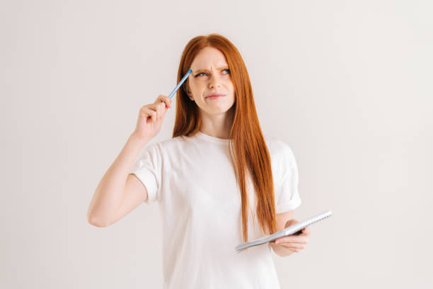 Studio portrait of pondering redhead young woman scratches head with pencil, holding diary looking up, standing on white isolated background. Pretty female student making notes in paper notebook.