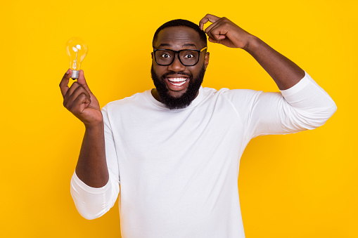 Photo of funky positive handsome guy hold light bulb scratching head toothy smile isolated on yellow color background.