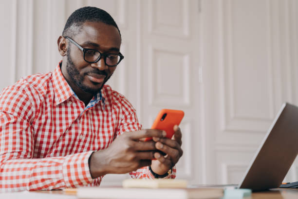 istock Young handsome african male executive in glasses sitting at office table and using mobile phone 1364195209