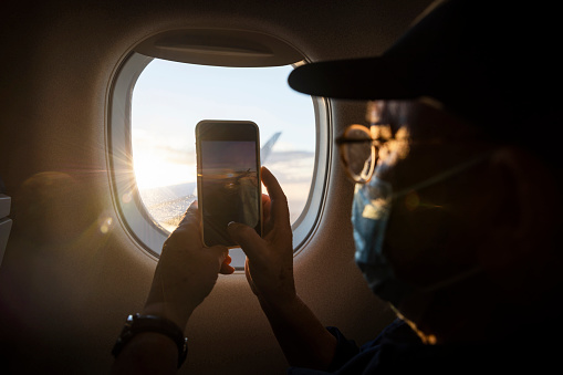 Passenger taking a picture on their phone out of an airplane window. They are wearing a protective face mask to help prevent the spread of Covid 19.