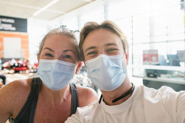 A female and male friend taking a selfie together while standing inside an airport terminal, ready to catch a flight home from France. They are wearing protective face masks, to reduce the spread of COVID-19.